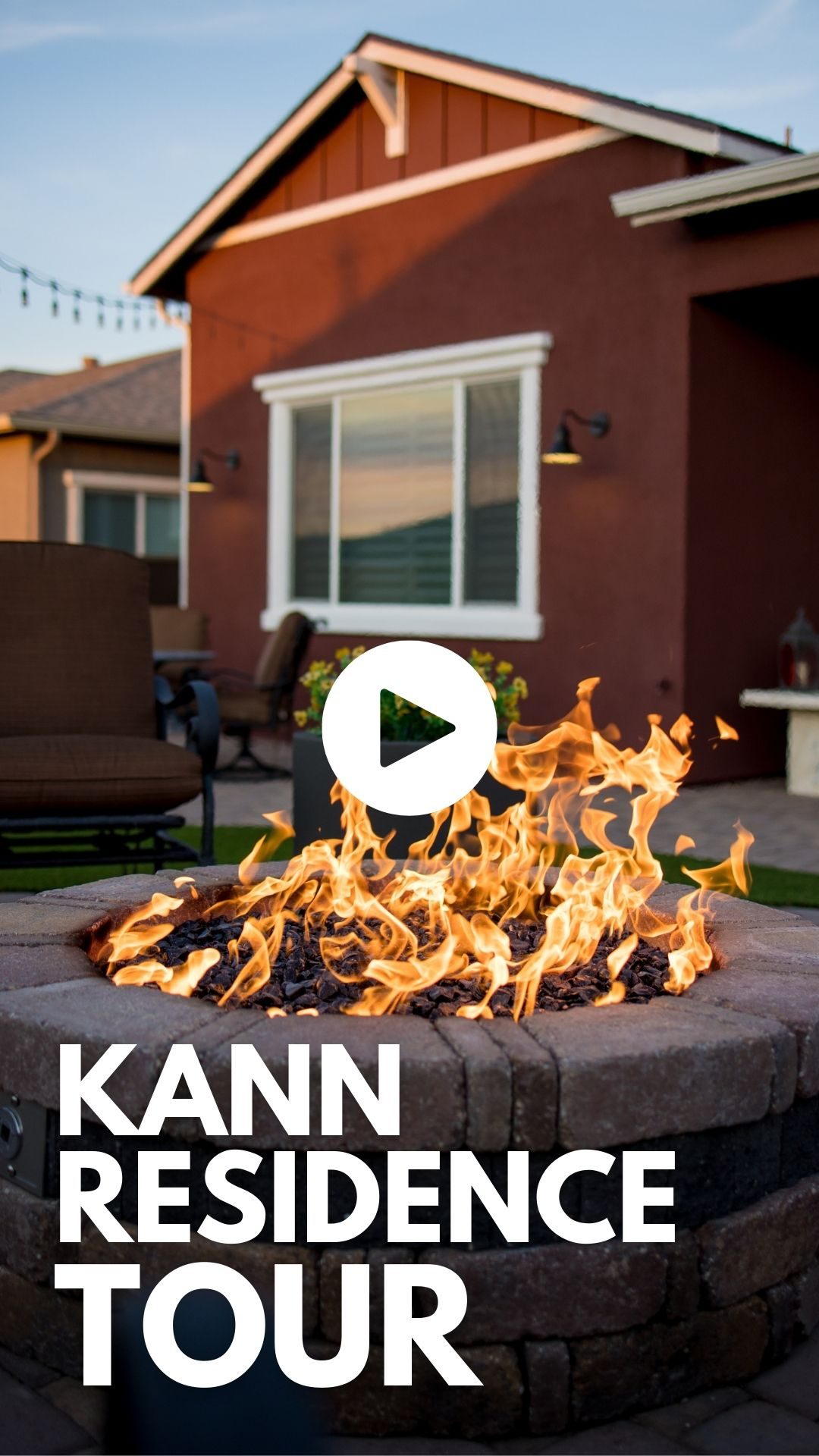 A cozy outdoor fire pit with bright flames in the foreground, surrounded by expertly laid pavers. In the background, a house with large windows and patio furniture is visible. Overlaid text reads "KANN RESIDENCE TOUR" with a play button icon above it.