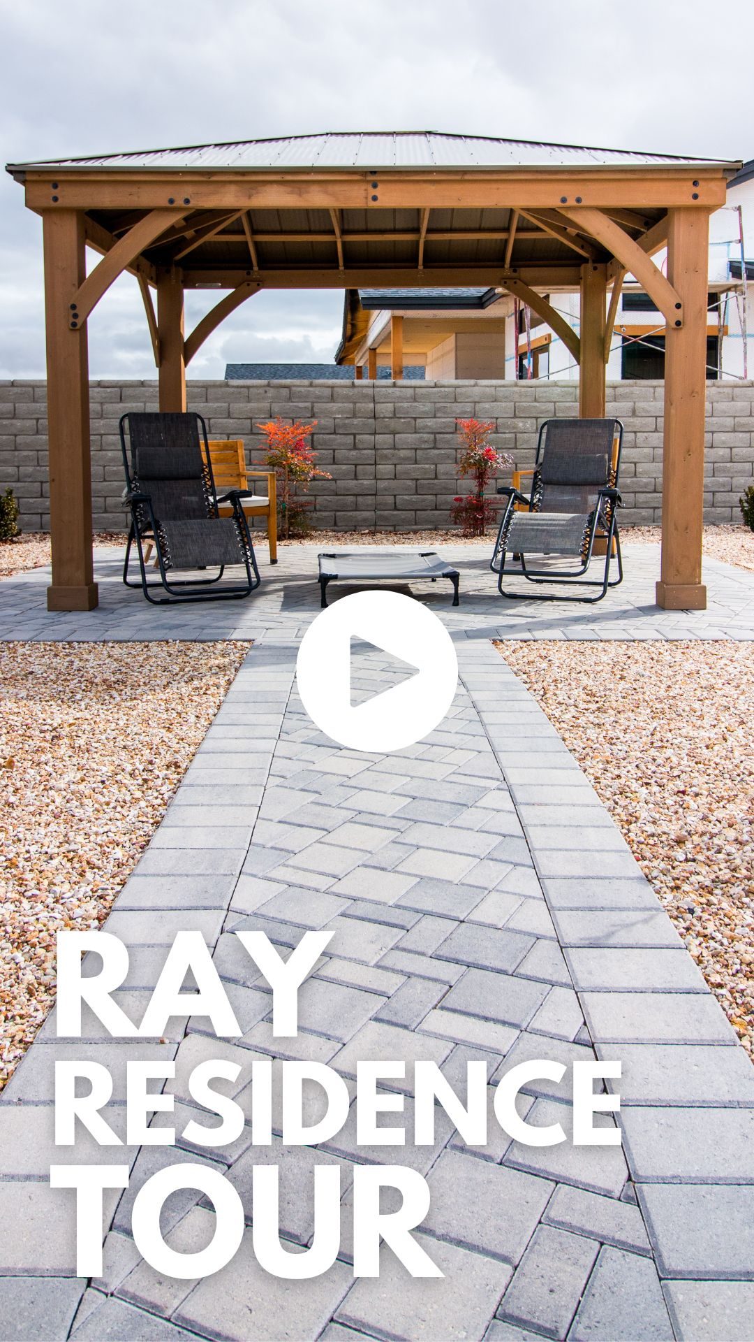 A photo of an outdoor patio area featuring a wooden pergola with a table and two lounge chairs underneath. The patio is surrounded by a stone pathway and gravel. Text overlays the bottom of the image, reading "Ray Residence Tour," with a play button icon in the center to start the virtual tour.