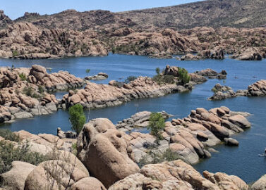 A scenic view of a lake surrounded by rocky formations and sparse greenery. A person on a paddleboard is on the calm water. The landscape is under a clear blue sky, creating a tranquil ambiance.