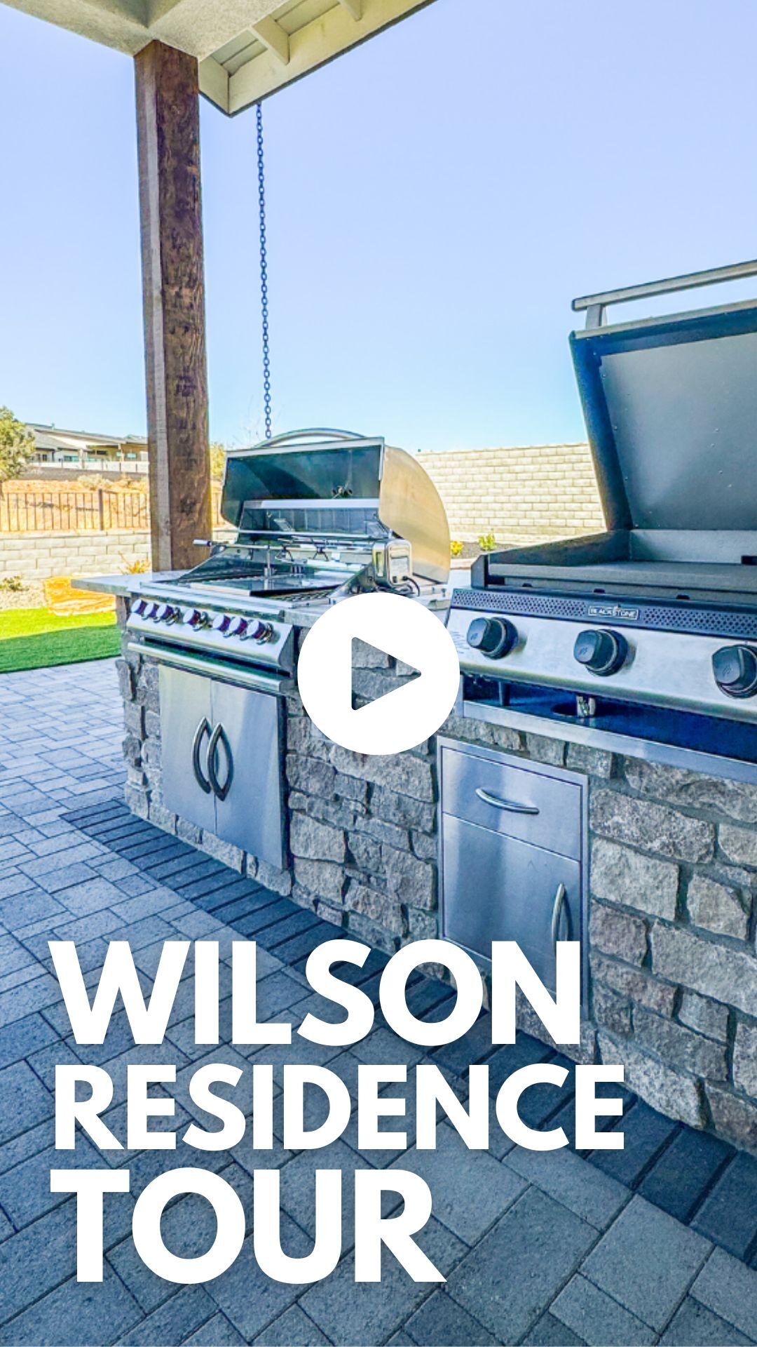 Outdoor kitchen with stainless steel grill and stone countertop, featuring a play button overlay, promoting the Wilson Residence Tour for Yavapai Landscaping's outdoor living space designs.