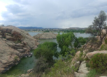 A scenic view of a lake surrounded by rocky cliffs and greenery. The sky is cloudy, and distant hills are visible in the background.
