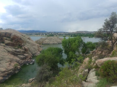A scenic view of a lake surrounded by rocky cliffs and greenery. The sky is cloudy, and distant hills are visible in the background.