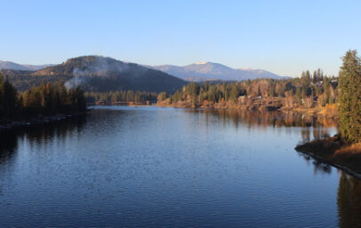 A serene lake stretches into the distance, bordered by forested hills under a clear blue sky. Sunlight gently illuminates the Chino Valley landscape, creating a peaceful and natural scene.