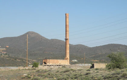 A tall, narrow brick chimney stands next to a small ruined building amidst the grassy landscape of Dewey-Humboldt, with distant hills under a clear sky. Power lines run across the scene.