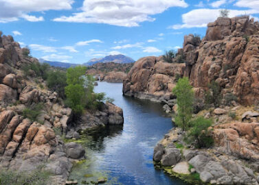 A scenic landscape of a narrow river flanked by rugged, rocky cliffs. Sparse vegetation dots the rocks, and the distant horizon features a mountain under a partly cloudy blue sky.
