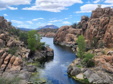A scenic landscape of a narrow river flanked by rugged, rocky cliffs. Sparse vegetation dots the rocks, and the distant horizon features a mountain under a partly cloudy blue sky.